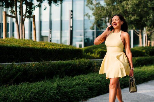 Smiling woman walking outdoors, holding a smartphone and a handbag, communicating in a modern urban park setting