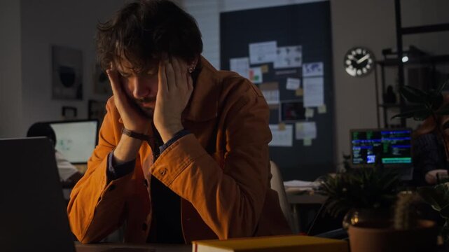 Young adult Caucasian man working late on laptop in office, rubbing temples and showing signs of fatigue and headache. Footage is ideal for corporate burnout and mental health at work concepts
