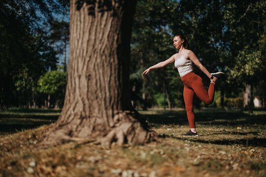 Woman performing a standing quad stretch in a sunny park to warm up for exercise. Outdoor fitness scene showing a casual workout and stretching routine in nature.