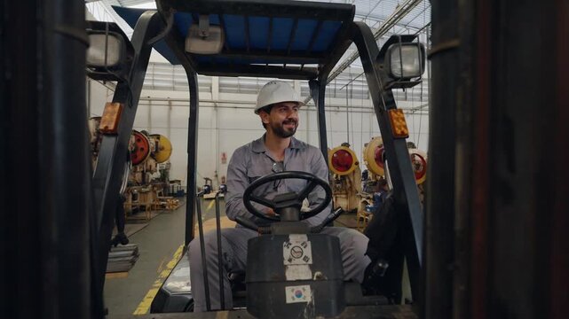 Man wears hard hat, grips forklift controls. Forklift operator rests hand on steering wheel. Factory background holds tools and storage racks. Determined look shows focus and readiness