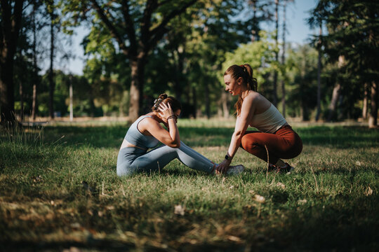 A personal trainer coaches a woman doing sit-ups during an outdoor fitness session in a park. Trainer holds the client's feet while she performs abdominal exercises on the grass.