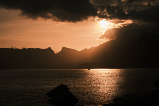 Stunning golden hour scene with sunbeams breaking through dark clouds, illuminating the tranquil ocean surface. Majestic mountains and a small boat are silhouetted, portraying nature's grandeur.