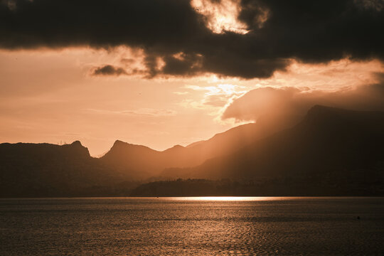 Breathtaking sunset over a tranquil lake, framed by majestic mountains and dramatic clouds. Golden light beams pierce through, creating a serene and powerful natural landscape.