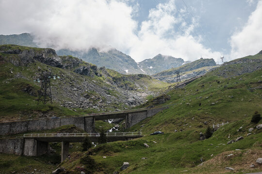 Majestic mountain pass with rugged green slopes and dramatic cloudy skies. Concrete bridges and power lines wind through the vast landscape, showcasing nature's grandeur meeting human engineering.
