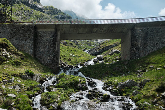 A sturdy concrete and stone bridge crosses a vibrant mountain stream cascading over rocks, surrounded by lush green slopes under a cloudy sky. Nature meets engineering in this scenic alpine landscape.