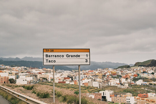 Road sign pointing to Barranco Grande and Taco, overlooking a vibrant hillside town with colorful houses, rolling hills, and a dramatic cloudy sky. A travel scene hinting at exploration and discovery.