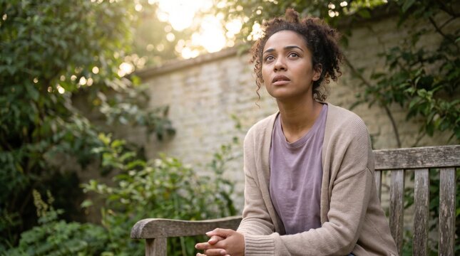 Young Black woman sits on a garden bench in soft morning sun, pausing with an alert reflective gaze as she uses outdoor light and fresh air to recover from an unsettled sleep pattern and regain calm