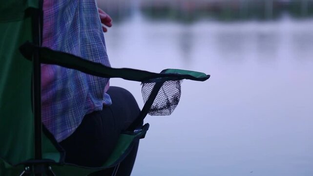 A fishing rod rests on a green holder near calm lake water. An angler wearing a plaid shirt reaches toward the reel with an outstretched hand, fingers moving to grip the handle.