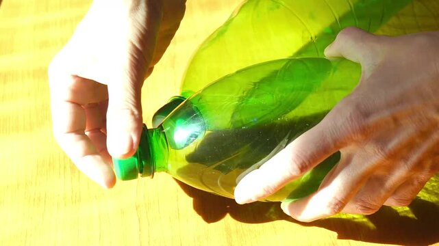 Flat lay close-up of hands unscrewing green plastic bottle cap on sunlit wooden surface. Shows household material sorting practice, recycling preparation, and responsible waste management habits.