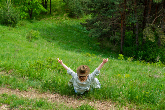 Blonde woman in green hat sitting on grass raising arms towards forest. Connection with nature, mental peace, outdoor recreation, and eco lifestyle concept.