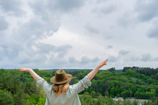 Blonde woman in green hat opening arms wide towards vast forest landscape. Harmony with nature, ultimate freedom, eco tourism, and outdoor adventure concept.