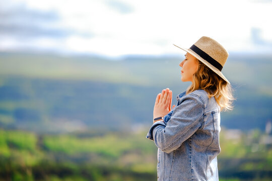 Calm woman in straw hat meditates with hands in prayer pose against serene mountain landscape. Idea of zen, mental health and inner balance in nature.