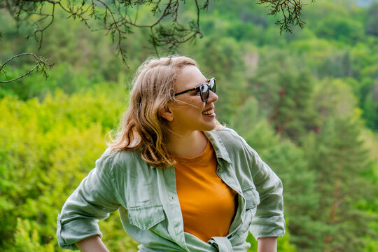 Cheerful blonde woman in sunglasses and light green shirt smiling outdoors in forest. Summer vacation, positive emotion, and active lifestyle concept.