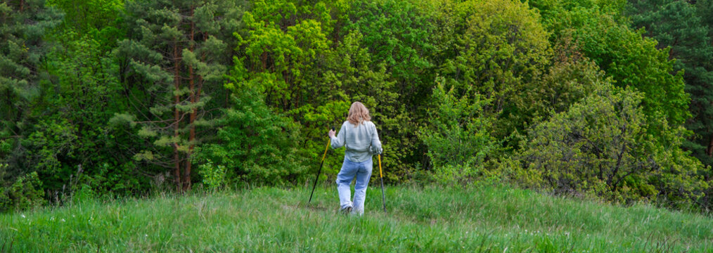 Active woman doing nordic walking in green meadow near forest from behind. Nordic walking, outdoor fitness, active longevity, and healthy lifestyle concept.