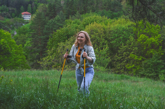 Smiling blonde woman doing nordic walking in green grassy field near forest. Healthy lifestyle, outdoor exercise, active recreation, and summer travel concept.