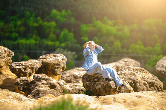 Smiling blonde woman in denim outfit sitting on ancient rocks in mountains. Summer travel, outdoor relaxation, mountain scenery, and freedom concept.