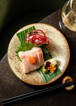 Top-down view of fresh white fish sashimi slices topped with orange tobiko roe, beautifully arranged on a speckled ceramic plate with shiso leaf, beet curls, pickled ginger, and edible flowers.