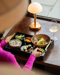 Over-the-shoulder high-angle shot of a woman in a pink sweater using chopsticks to eat sushi from a Japanese bento set served on a tray with green curry, edamame, rice, and wine. © Simone Oppes