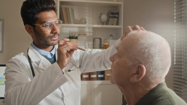 Indian young adult doctor examining eyes of Caucasian senior man with penlight in clinic. Footage ideal for geriatric healthcare services, medical checkups and senior care advertising