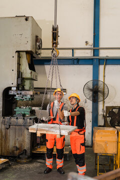 Factory workers guiding overhead crane lifting wrapped metal pallet near power press