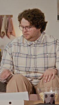 Vertical portrait of Caucasian young man with glasses pointing TV remote at camera, switching channels on sofa. Clip is perfect for streaming service or home entertainment ads, vertical