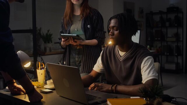Handheld shot of multiethnic young adult colleagues collaborating late at night in office, using laptop and tablet. For corporate teamwork, startup culture, and business deadline concepts