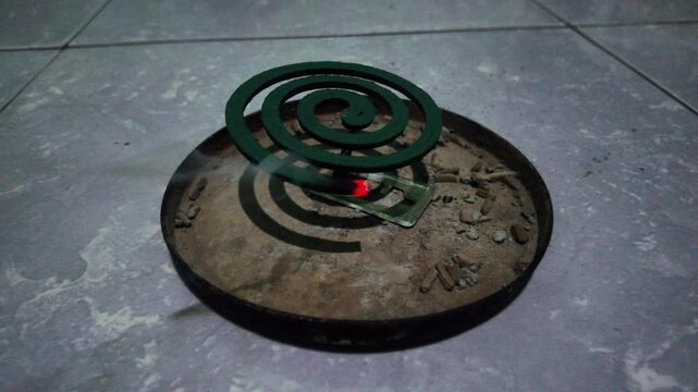 Burning green mosquito repellent coil emitting smoke on a metal tray with ash on the floor
