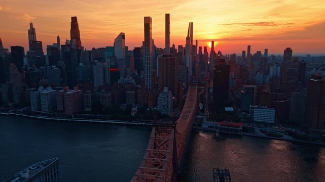 Flyover the Queensboro Bridge in New York City. Metropolis skyscrapers against the orange sky with beautiful setting sun.