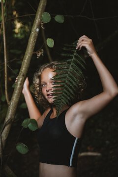 Teenage girl stands in a forest, playfully holding a fern leaf near her face, blending with the natural surroundings. Her expression conveys a sense of curiosity and engagement with nature.