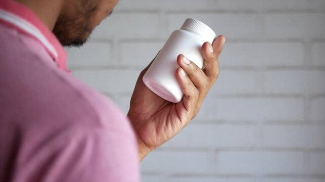 A man in a pink shirt is looking at a medicine bottle in his hand with a white brick wall in the background, a static over the shoulder shot