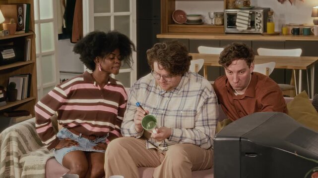 Diverse young adults sitting together, watching television and sharing drink from mug. Perfect for marketing campaigns focusing on friendship, home entertainment, or retro lifestyle