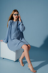 Beautiful fashionable woman wearing trendy blue color pleated short dress, sunglasses, silver metallic strap sandals. Full-length studio portrait. Copy, empty space for text © Victoria Fox