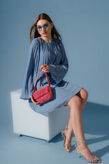 Beautiful fashionable woman wearing trendy blue color pleated short dress, sunglasses, silver metallic strap sandals, holding pink beaded bag. Full-length studio portrait. Copy, empty space for text © Victoria Fox