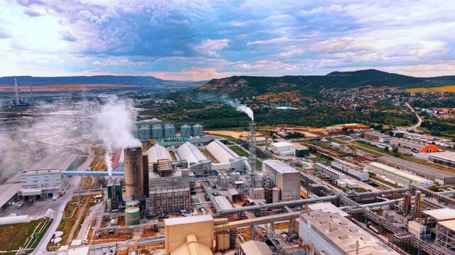 Industrial factory complex with smoking chimneys. White smoke rises from a manufacturing plant situated near a rural town and green hills under a cloudy sky.