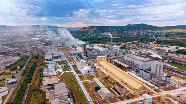 Industrial manufacturing plant with long warehouse buildings. Smokes rise from chimneys across a vast production area bounded by hills and a suburban settlement.