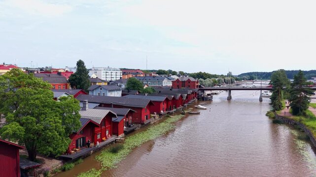 Low aerial view of Porvoo's iconic red wooden riverside warehouses with private decks along Porvoonjoki river, colorful old town houses, road bridge, and moored boats, Finland