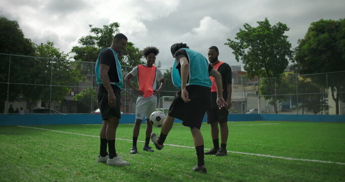 Young Hispanic man kicking soccer ball during casual game with friends, losing balance and falling as teammates laugh playfully, relaxed atmosphere of friendship and fun