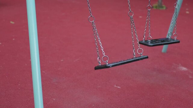 Two empty swings gently sway in a playground, with a vibrant red rubber surface and a turquoise frame, inviting children to play and enjoy outdoor activities