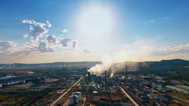 Massive industrial zone with smoking chimneys under a bright midday sun. A high angle drone view displays a factory area releasing white smoke against a clear back lit sky.