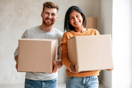 Happy young mixed race causasian and indian couple holding boxes in new home or apartment, real estate marketing