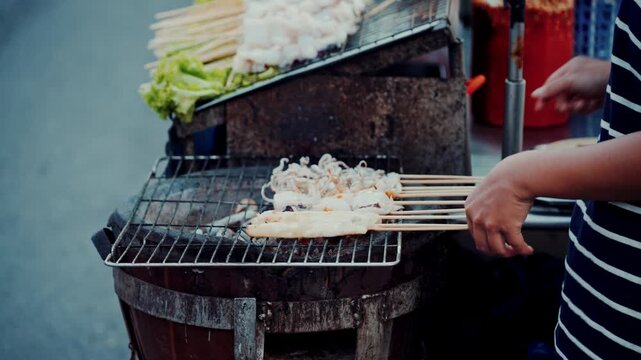 Close up of vendor grilling fresh squid skewers on hot charcoal stove in night market in Thailand.