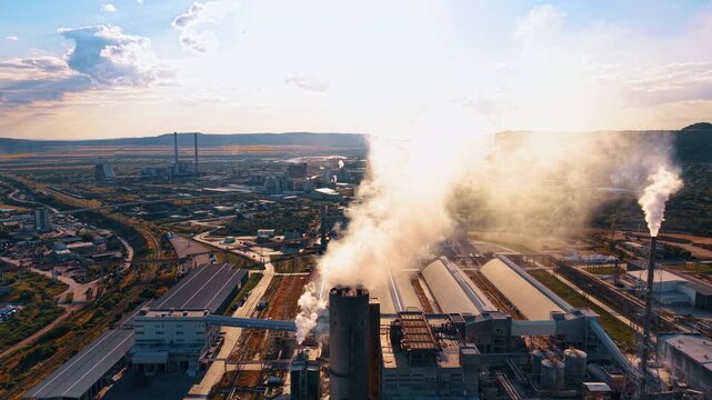 Thick white smoke billows from a large industrial tower at sunrise. An aerial shot reveals factory rooftops and tall stacks discharging emissions into a bright hazy atmosphere.