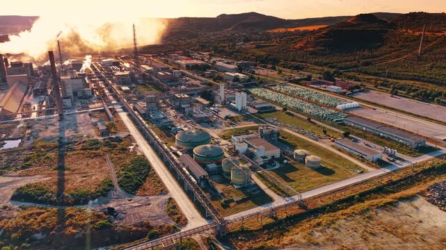 Industrial refinery plant with circular storage tanks during golden hour. A drone perspective shows green landscape fields surrounding oil processing facilities in warm evening light.