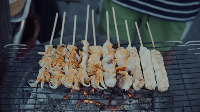 Close up of vendor grilling fresh squid skewers on hot charcoal stove in night market in Thailand.