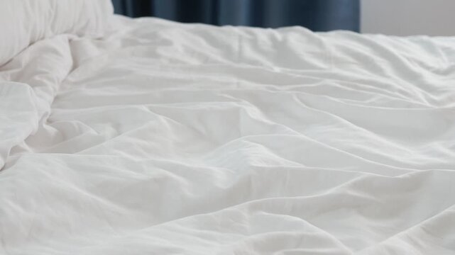 Slow motion shot of white wrinkled bed sheets on empty mattress in daylight bedroom interior with window curtains in background