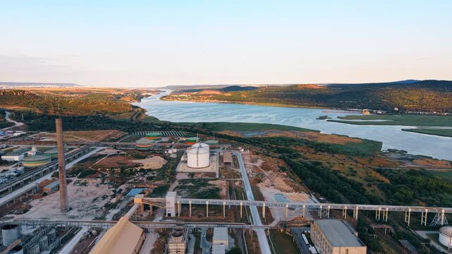 Aerial view of an industrial facility along a wide river.. Manufacturing buildings and storage tanks sit next to a large water body surrounded by green hills.