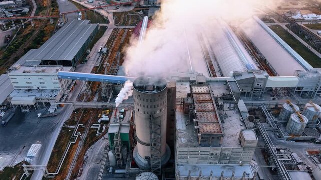High angle view of a factory chimney emitting white smoke.. Dense white smoke rises from a concrete tower over industrial roofs and piping networks.