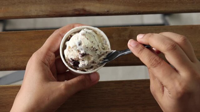 Top-down view of hands holding small cup of cookies and cream ice cream and scooping it with tiny black spoon on brown wooden slatted background