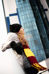 Low angle profile portrait of a thoughtful young woman in a striped Gryffindor style scarf and beanie, looking away against a modern glass skyscraper background. © Anatoliy Novikov