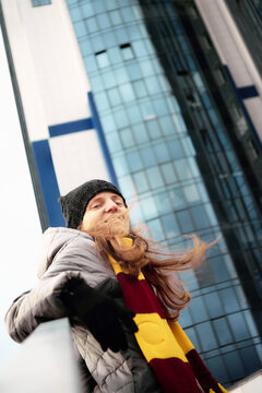 Low angle portrait of a smiling young woman with long windblown hair, wearing a striped Gryffindor style scarf, beanie, and winter jacket against a modern glass skyscraper.
